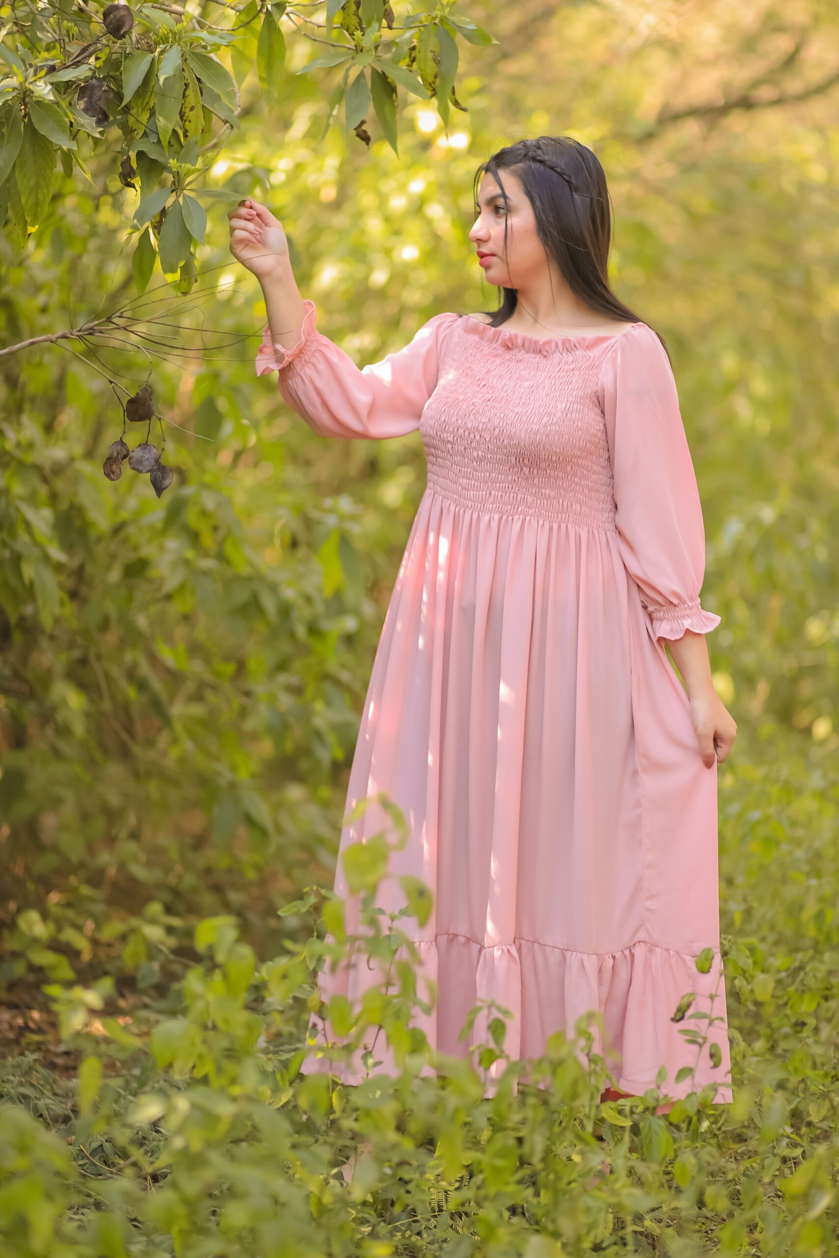 A woman wearing a pastel pink long-sleeve maxi dress stands in a lush green garden, gently touching tree leaves in a peaceful natural setting.