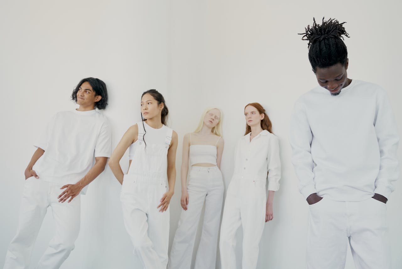 A diverse group of adults in white outfits posing against a white wall in a studio.