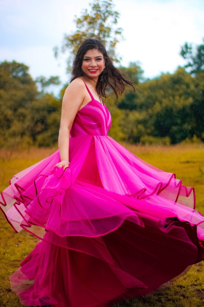Smiling young woman twirling in a vibrant pink layered tulle gown in an outdoor field.